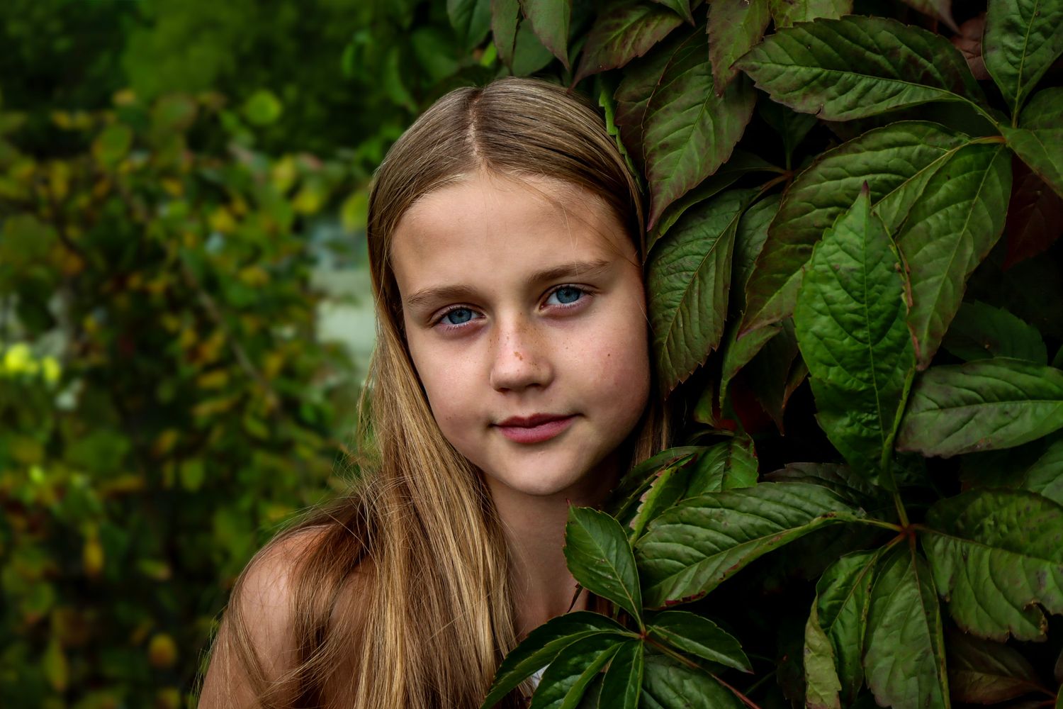 Nærbilde av jente med blondt hår fotografert mellom store grønne blader.