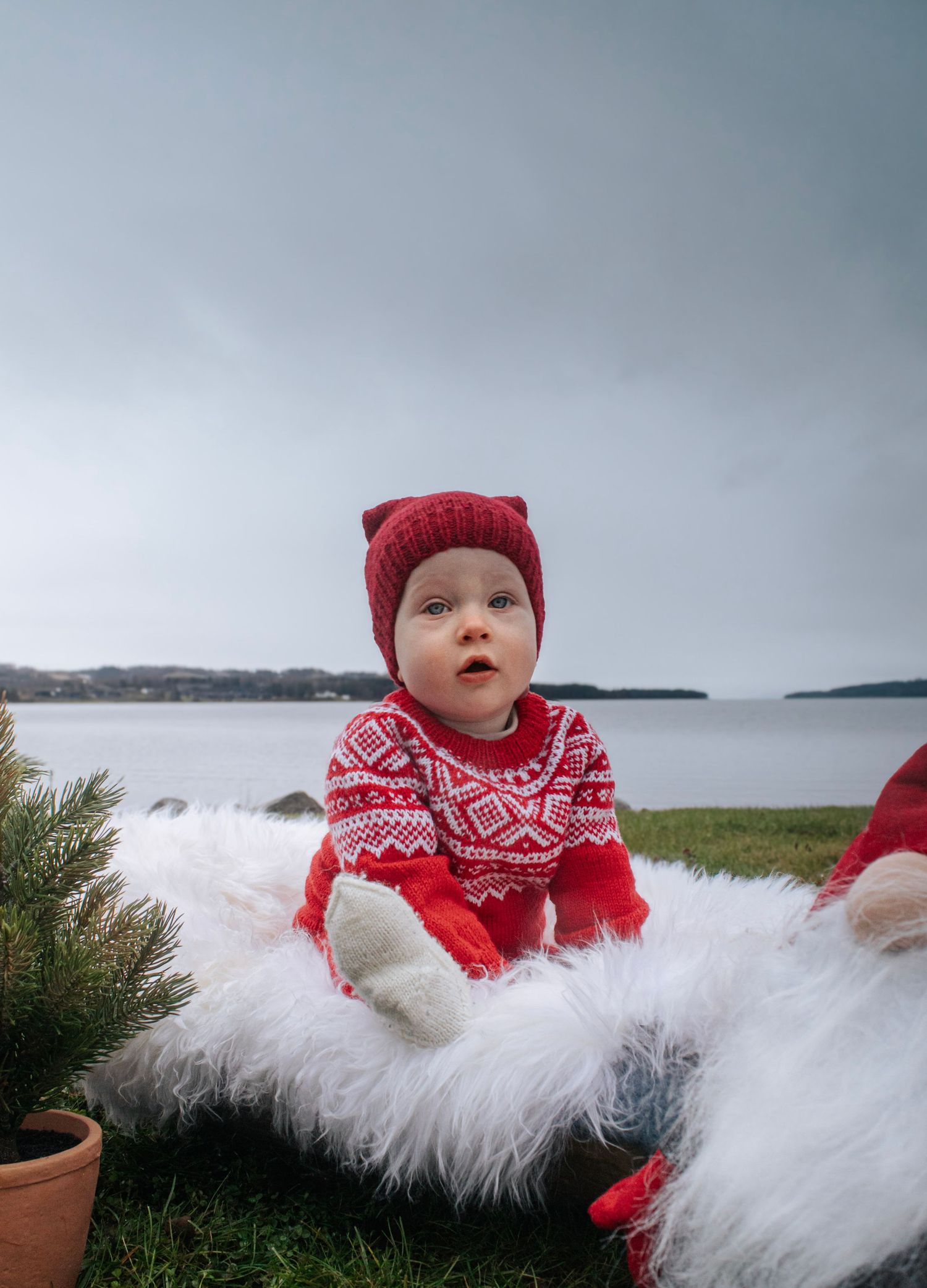 Baby i rød strikkegenser og nisselue sittende på hvitt skinn utendørs ved vannet.