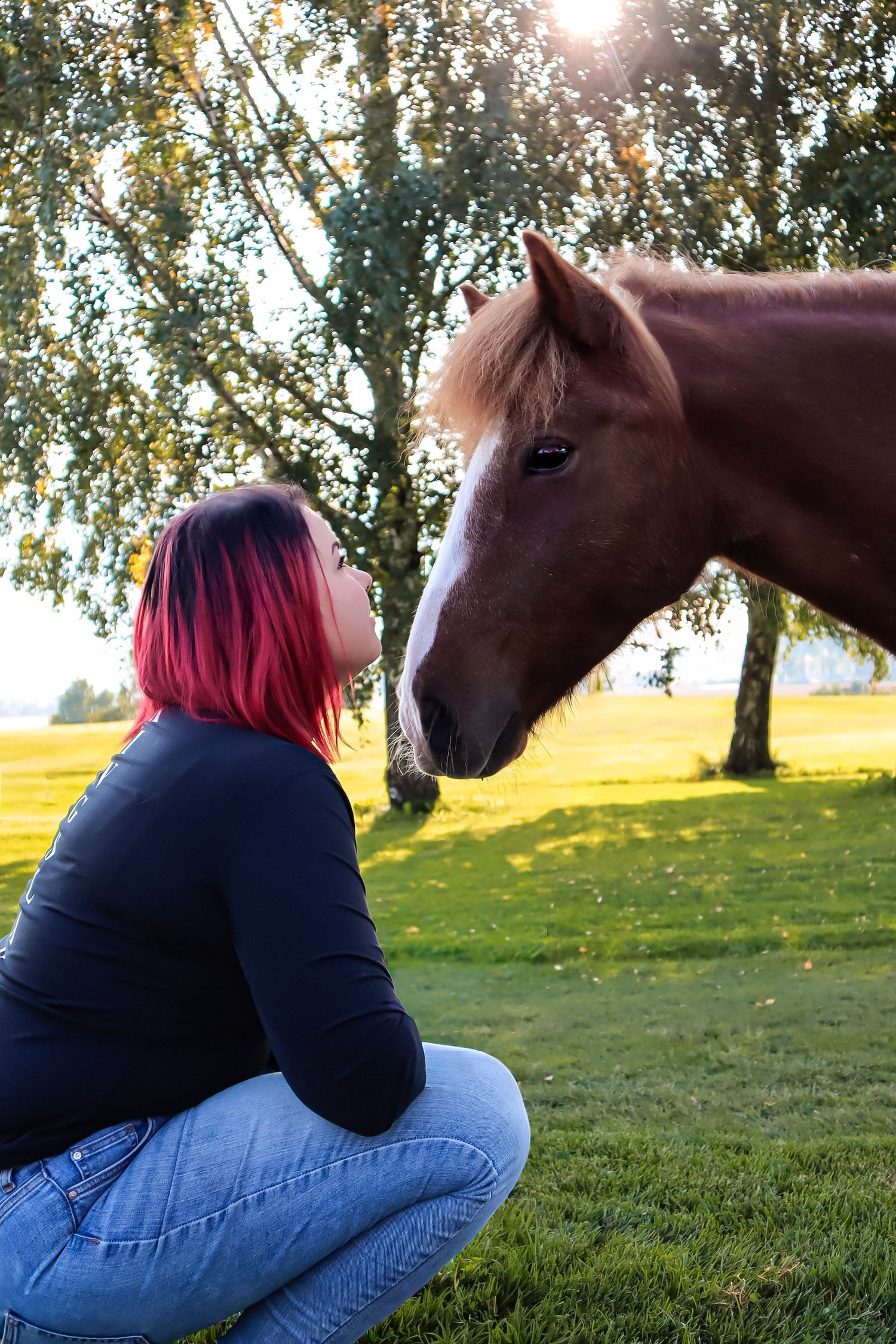 Kvinne med rødt hår sammen med en brun hest på et grønt beite i kveldssol.