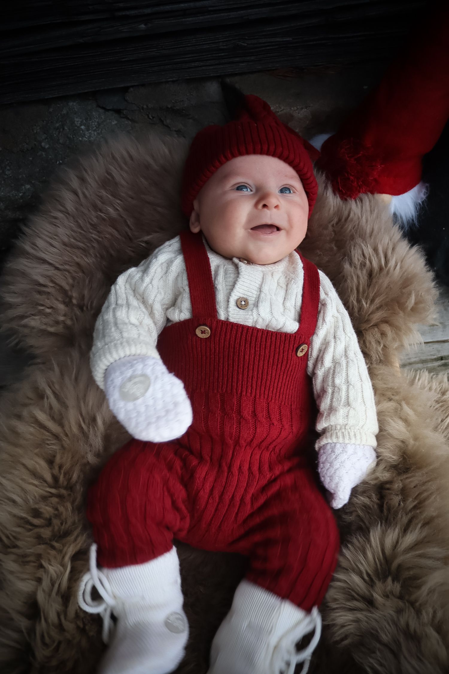 Smilende baby i rød strikkebukse og nisselue liggende på saueskinn.