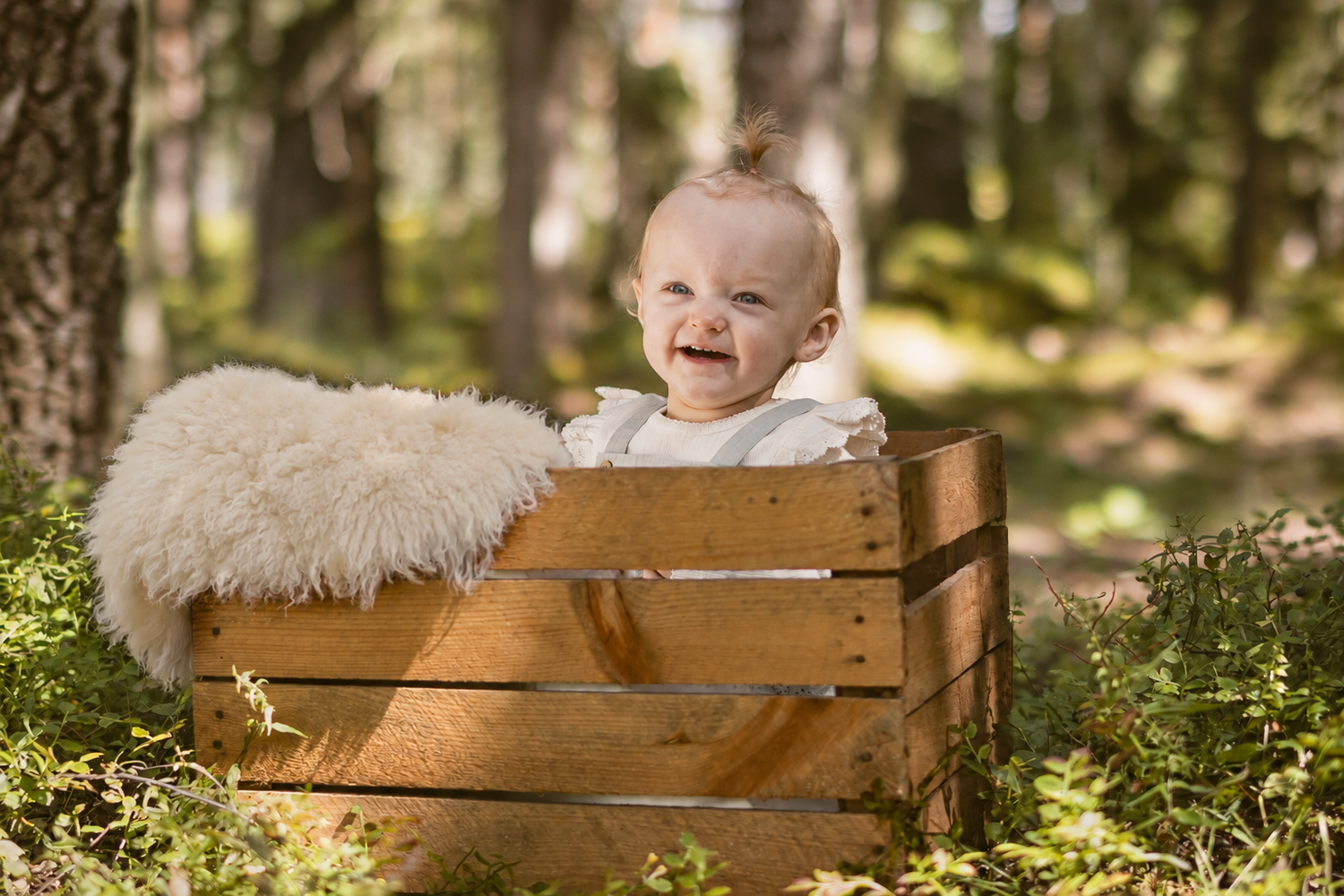 Leende baby sittende i en trekasse med hvitt teppe i skogen.