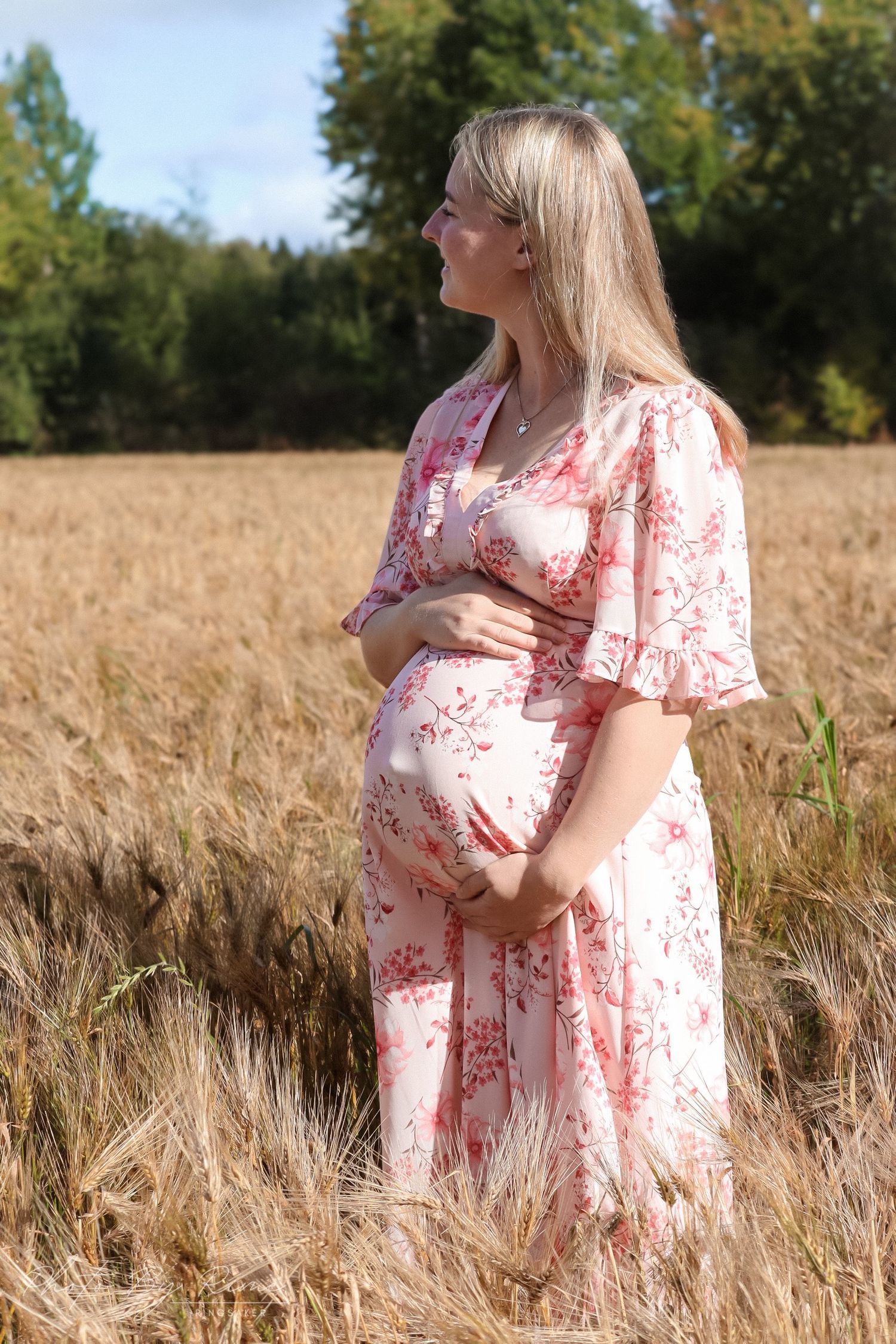 Gravid kvinne i rosa blomsterkjole fotografert i en kornåker.