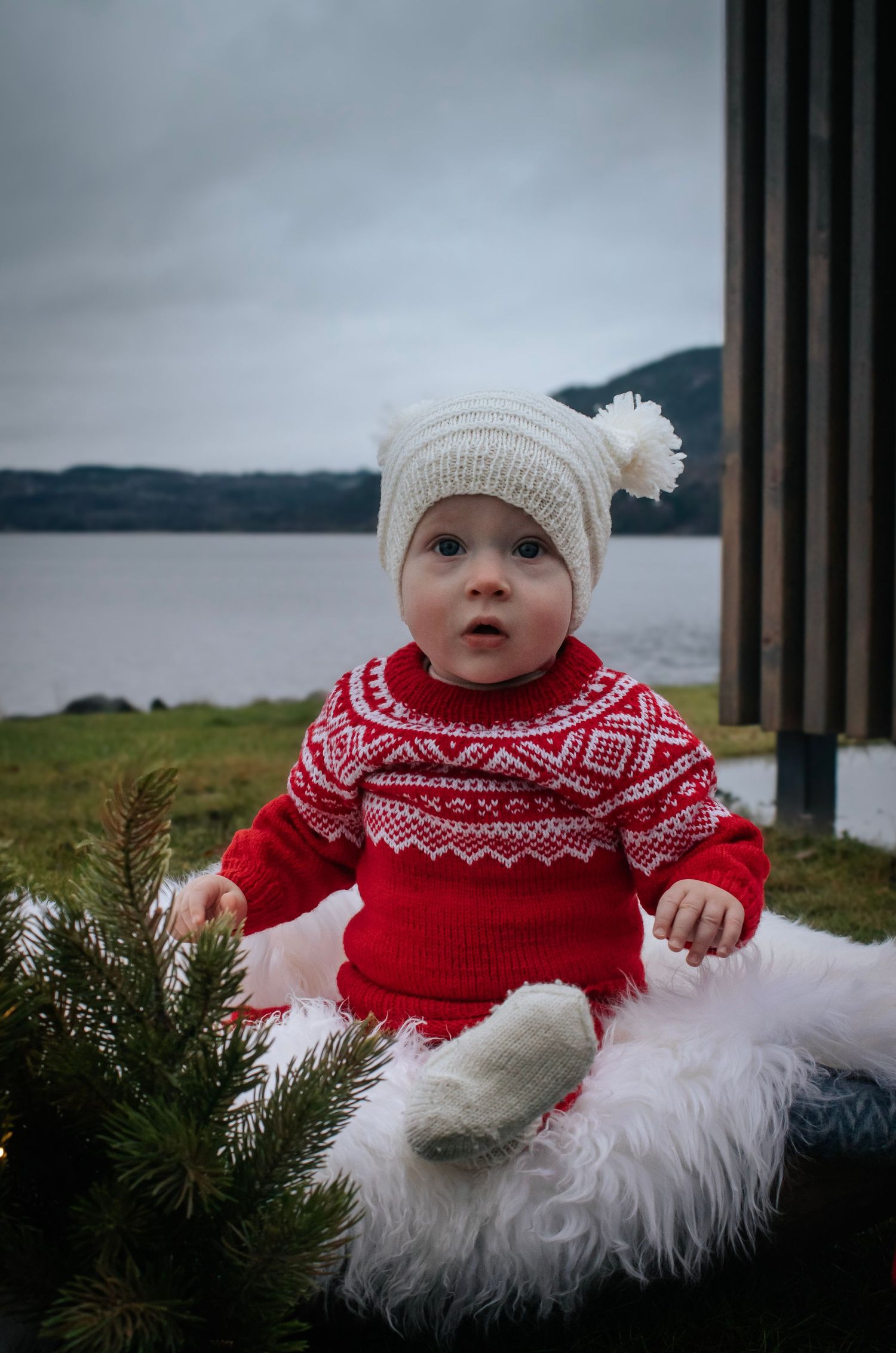 Baby i rød mønstret strikkegenser og hvit lue fotografert ved granbar ved vannet.