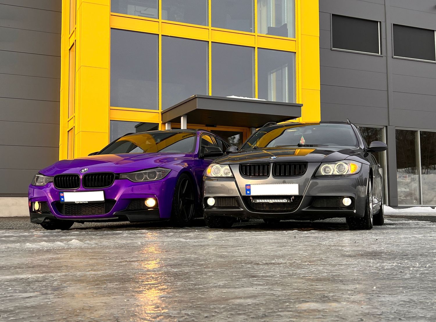 Lilla og mørkegrå BMW fotografert side om side foran en gul bygning.