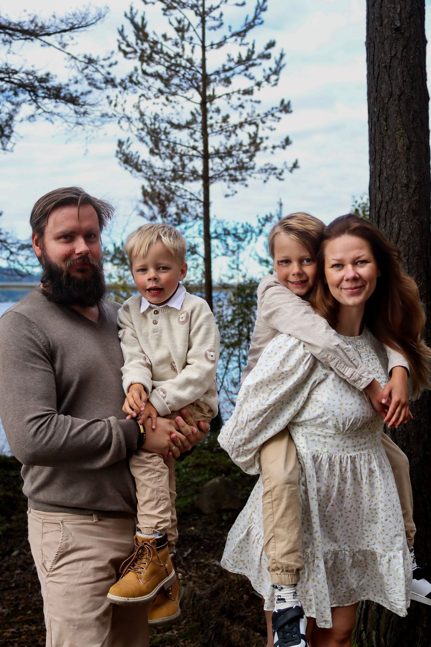 Familie på fire fotografert utendørs blant furutrær med Mjøsa i bakgrunnen.