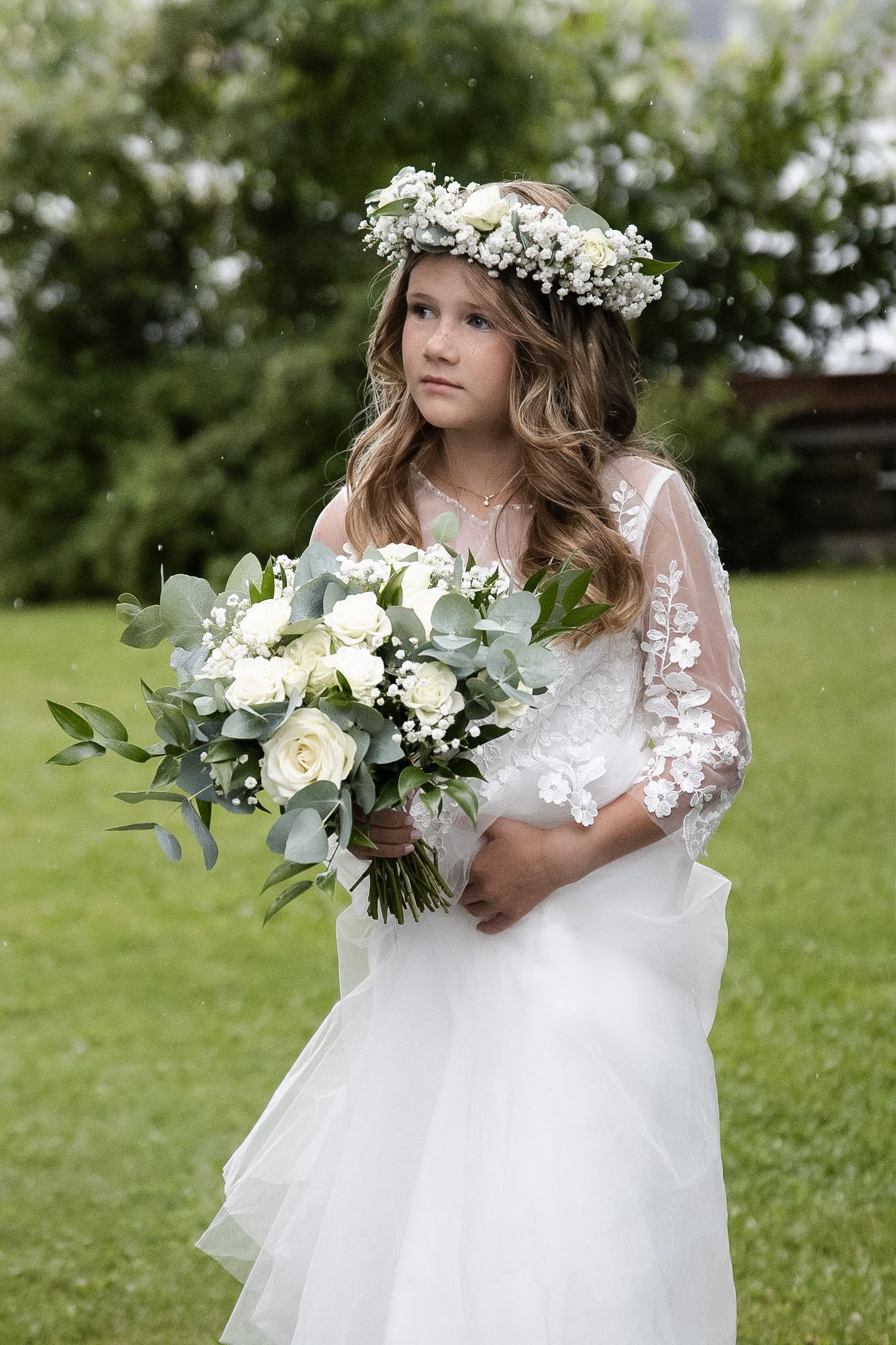 Brudepike i hvit blondekjole med blomsterkrans og bukett fotografert utendørs.