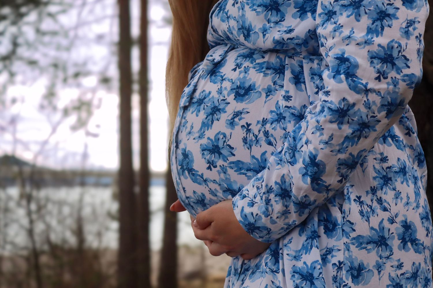 Nærbilde av gravid kvinne i blå blomstrete kjole med hendene rundt gravidmagen.