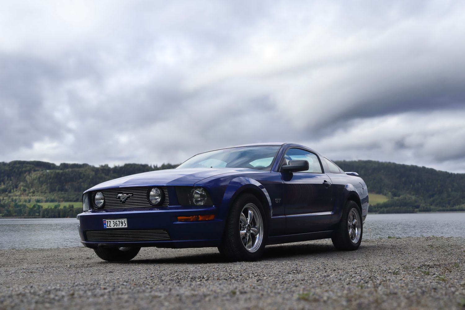 Mørkeblå Ford Mustang fotografert på grus ved Mjøsa med åser i bakgrunnen.