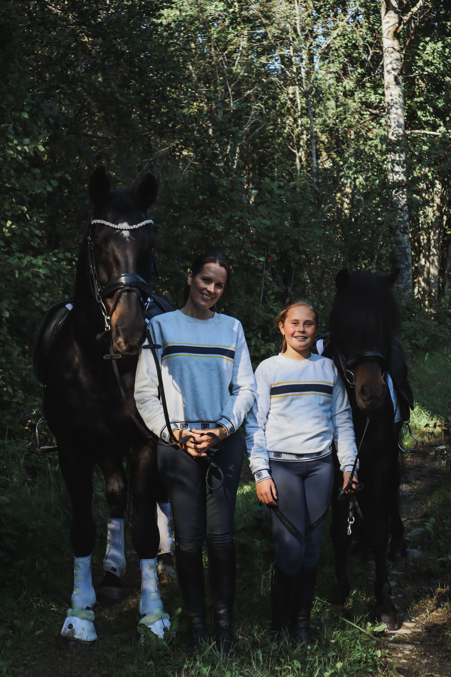 Mor og datter fotografert sammen med hver sin mørke hest i skogen.
