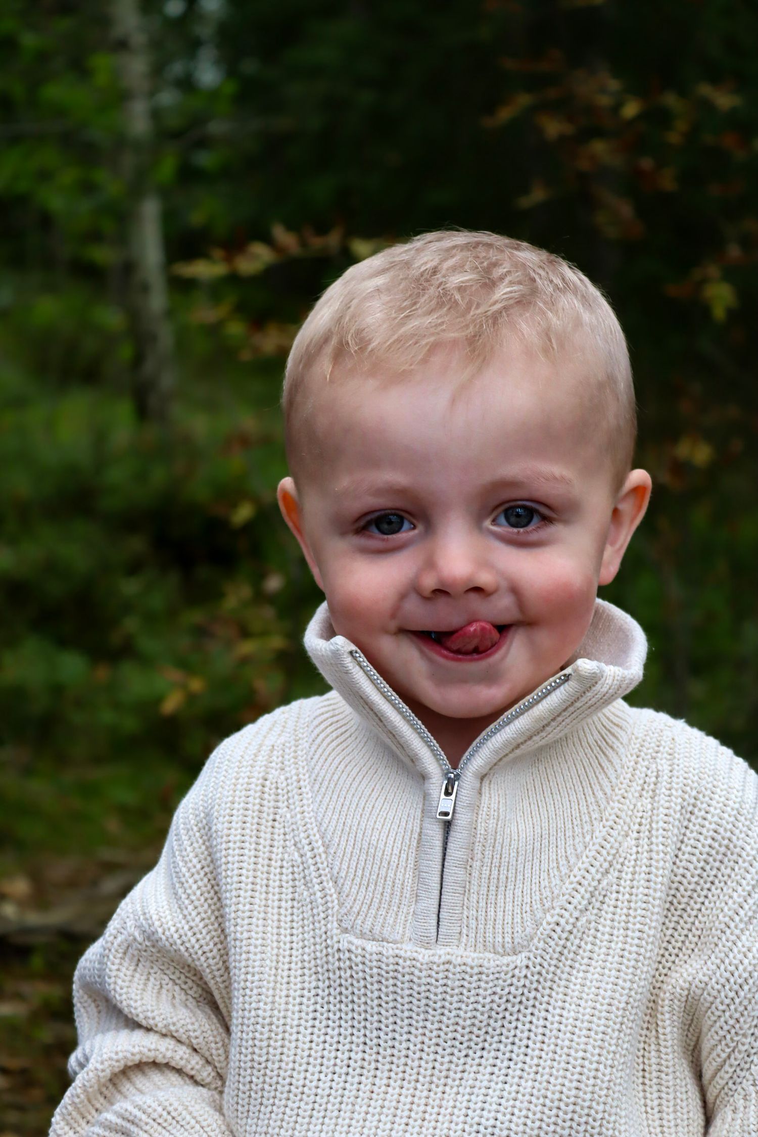 Liten gutt i lys genser fotografert i skogen med tungen ut.