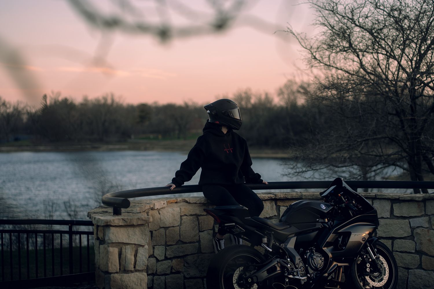 A motorcyclist in black gear and helmet leans against a stone wall beside a sport bike at dusk near a serene lake.