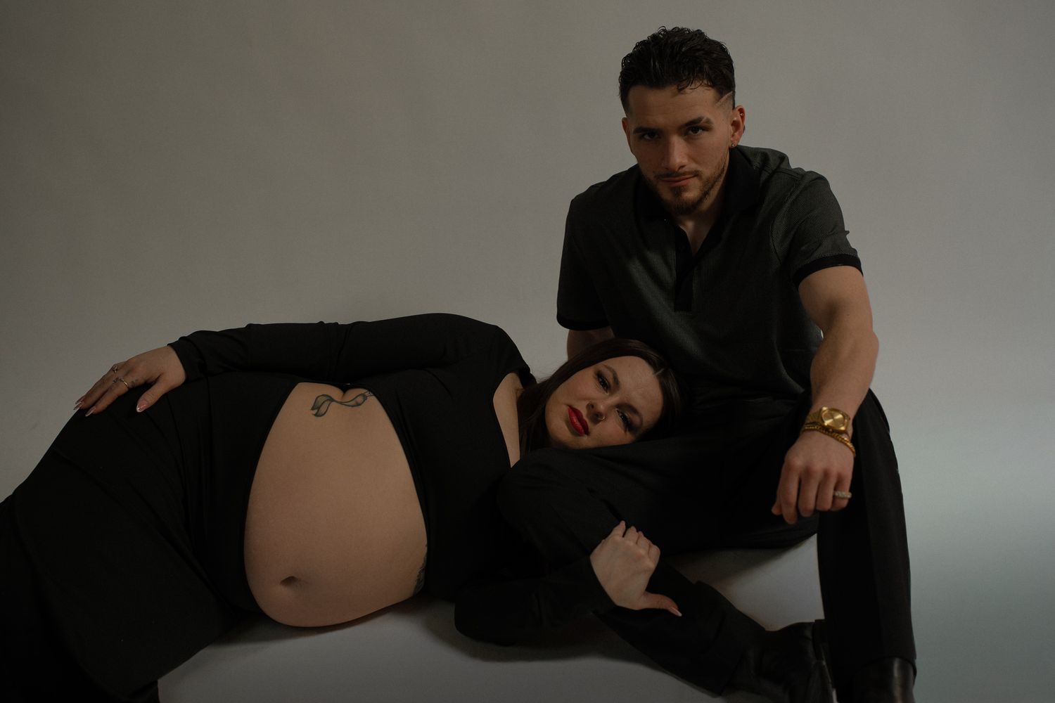 A maternity photoshoot featuring a couple in black clothing against a neutral backdrop.