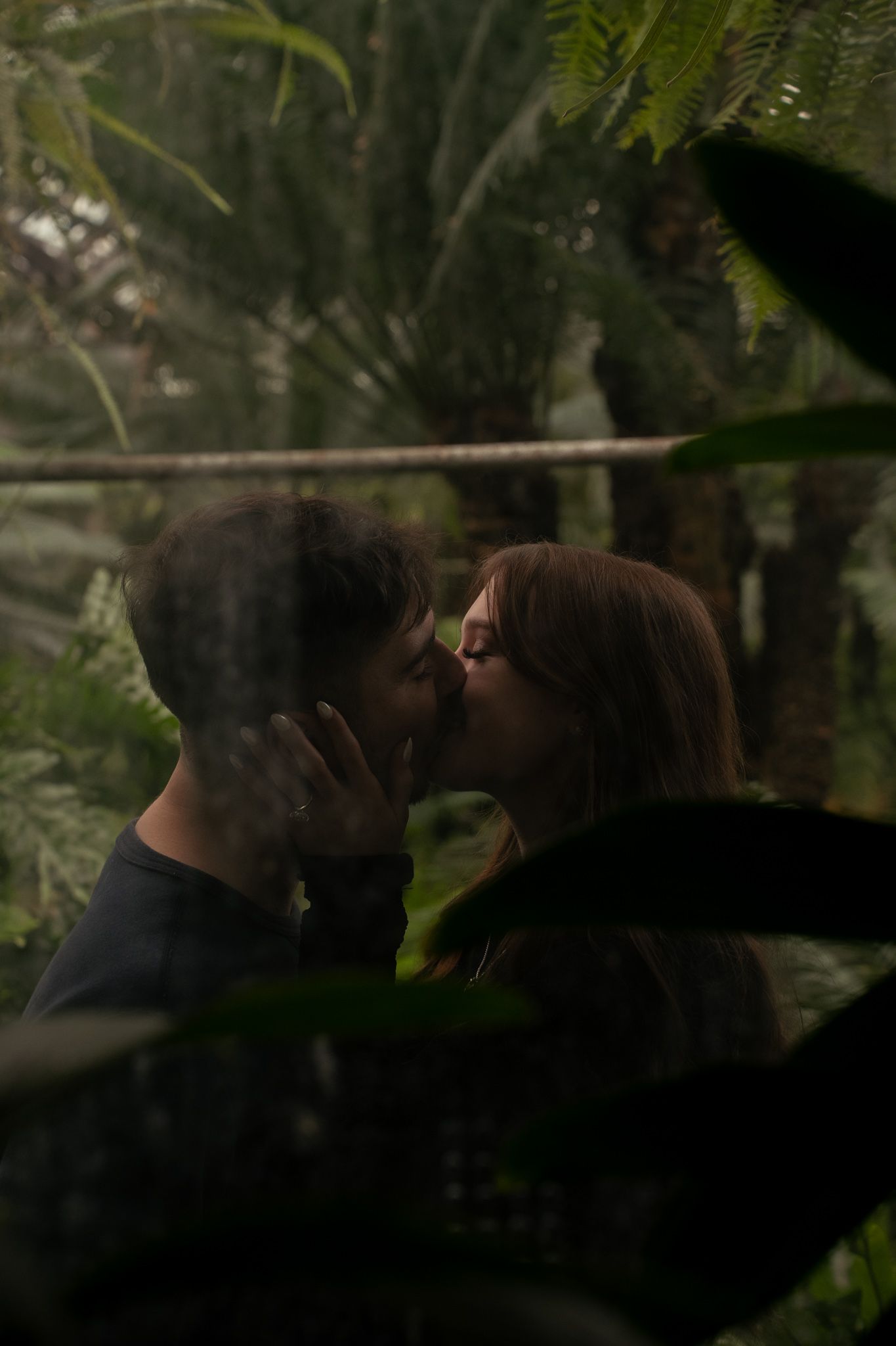 A couple shares a tender kiss in a dark, moody forest setting with green foliage in the background.