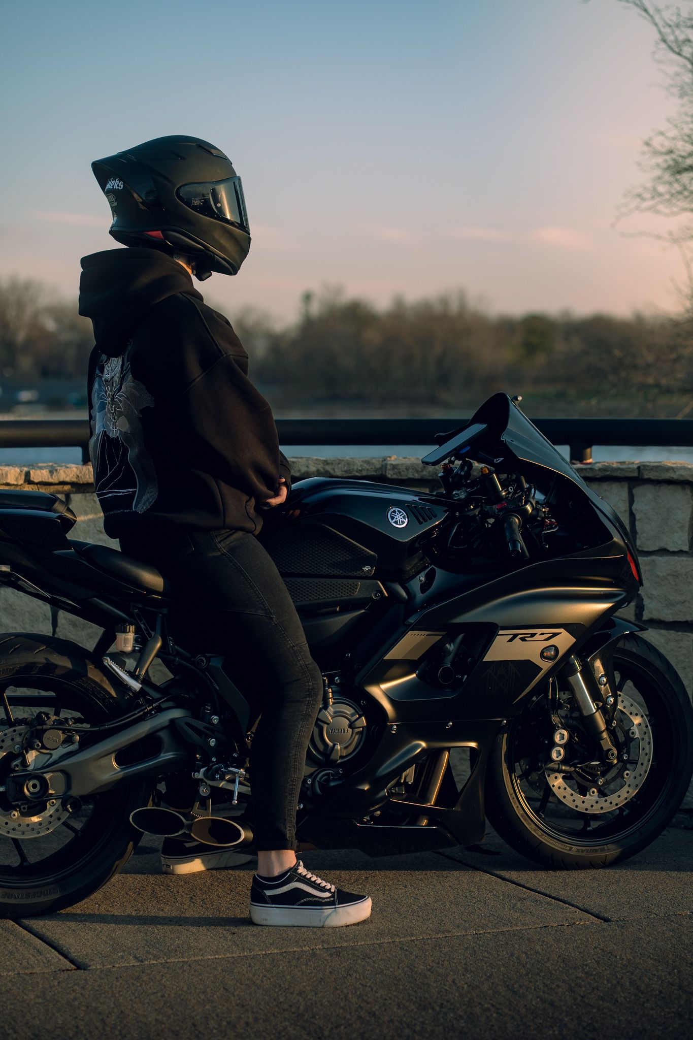 A rider in black gear and helmet stands beside a sleek black Yamaha R7 sport motorcycle at sunset.