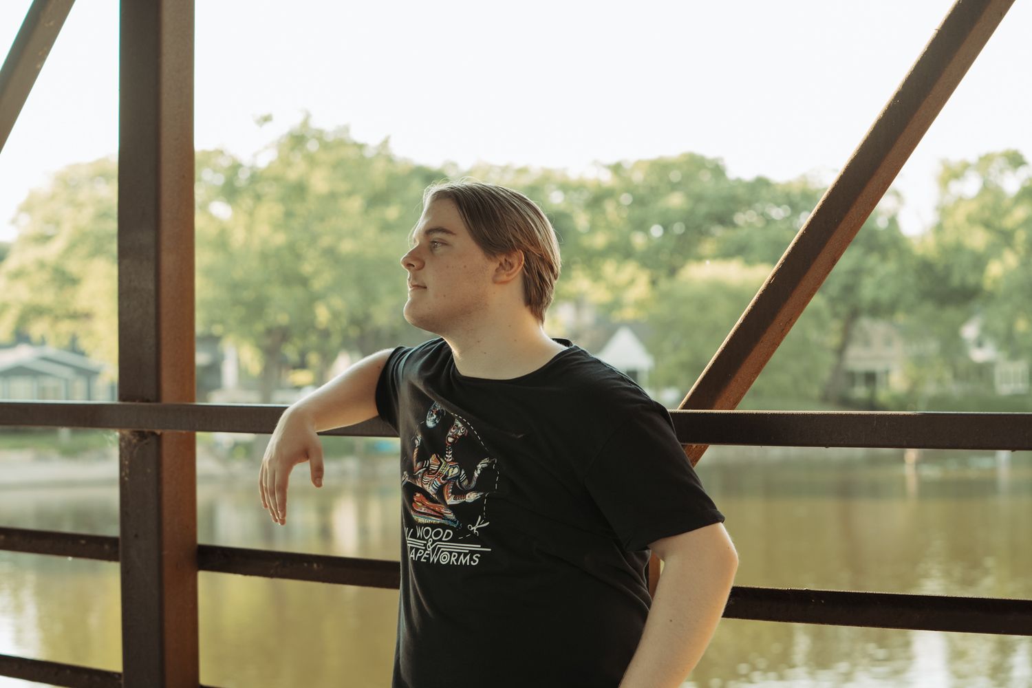Young person in black t-shirt leaning on bridge railing with river and trees in background.