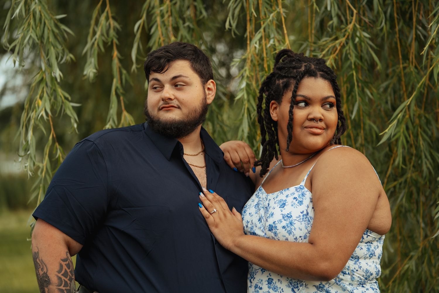 A couple poses under a weeping willow tree, man in navy shirt with tattoos, woman in floral sundress.
