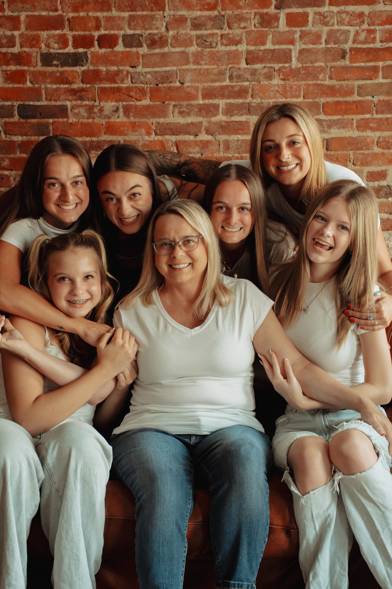 A cheerful woman with glasses poses with six girls on a couch against a brick wall, all wearing white tops.