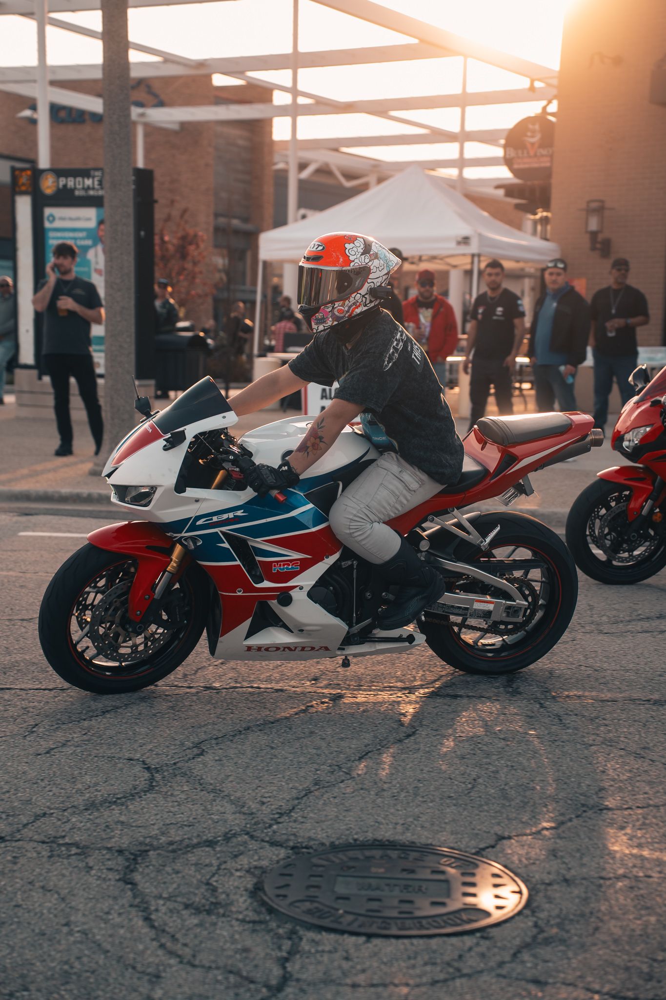 Rider on a Honda CBR sportbike at a motorcycle meet, wearing a helmet, with crowd and golden sunset in background.