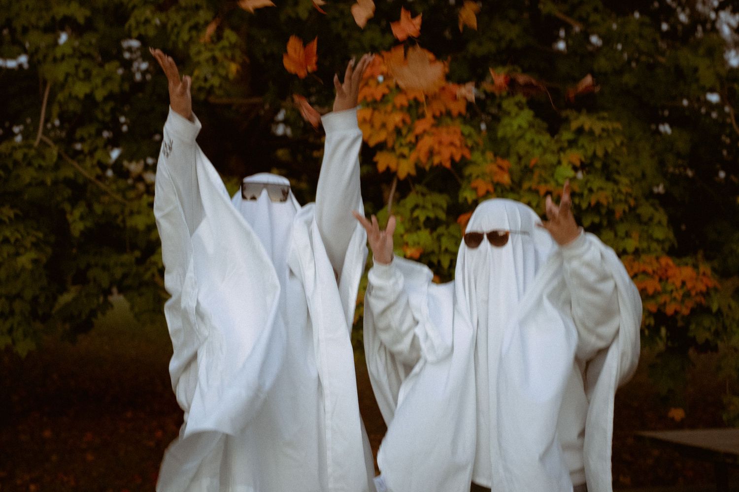 A group of people dressed as ghosts in white sheets pose together among fall foliage.
