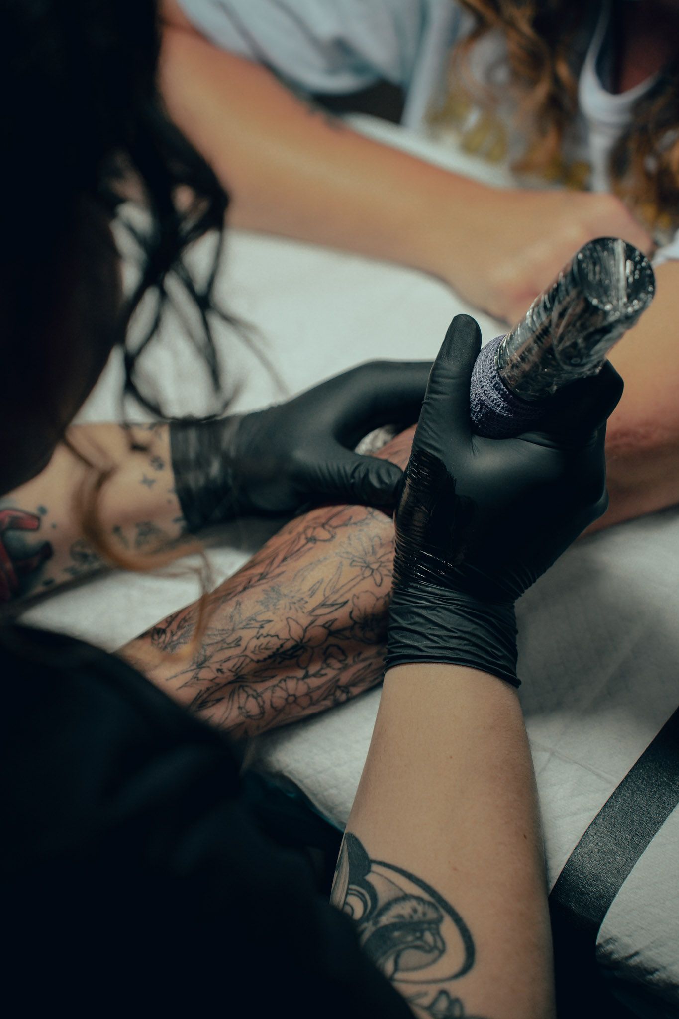 Tattoo artist wearing black gloves carefully tattooing a client's leg in a professional studio setting.