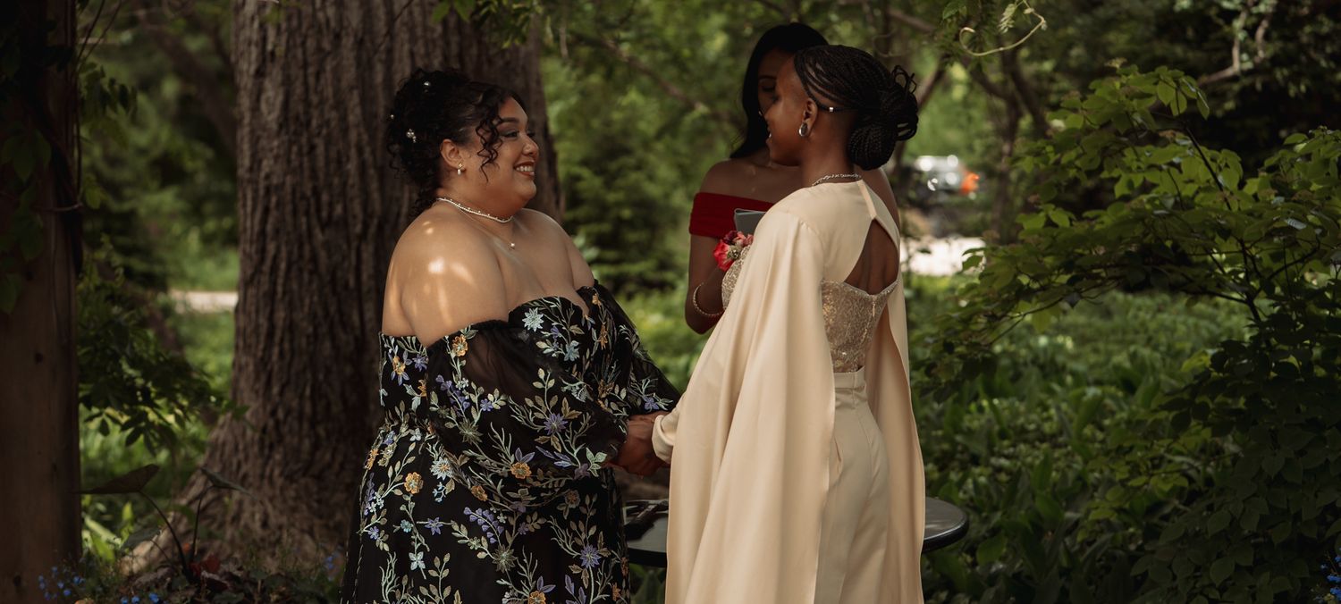 Two women in elegant dresses converse outdoors among lush green trees in a garden setting.