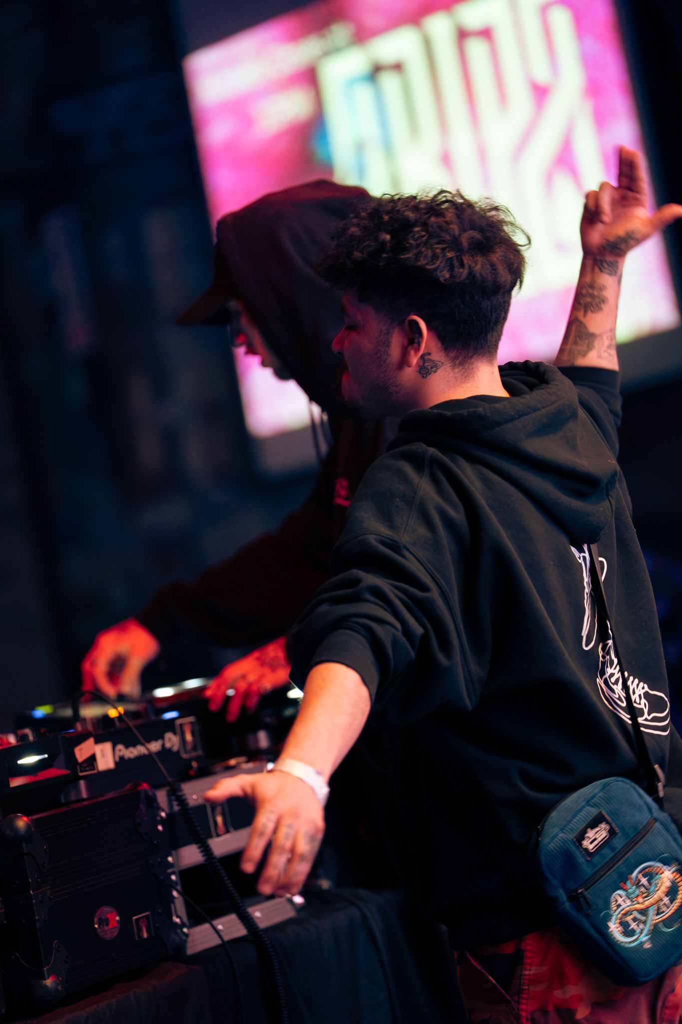 A tattooed DJ in a black hoodie and cap raises one hand while mixing music at a DJ booth under colorful stage lights.
