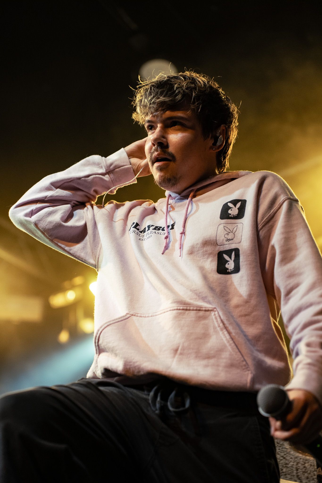 Person wearing a light pink hoodie with a logo sits against a dark background with dramatic lighting.