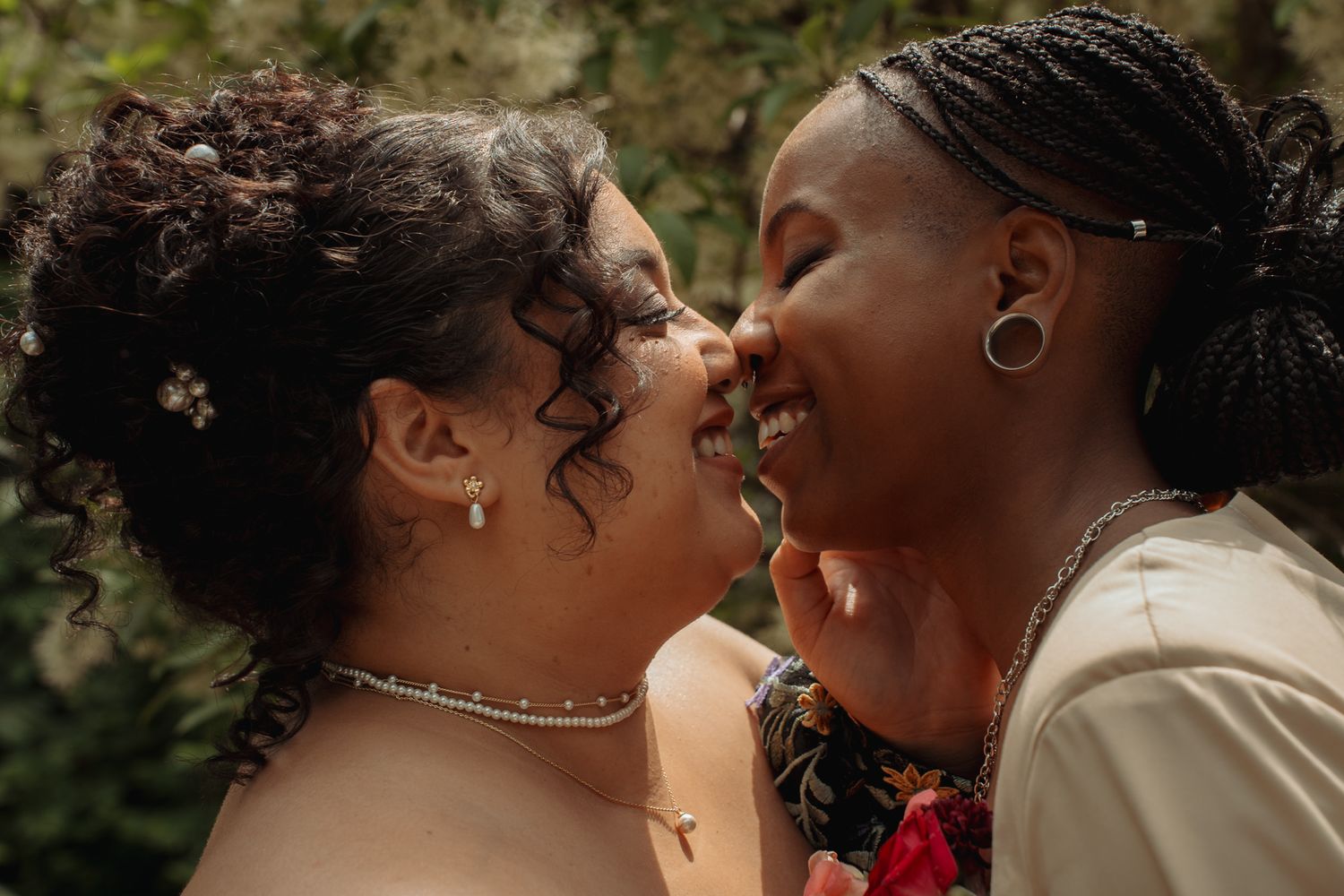 Two women smiling and sharing a joyful moment outdoors, wearing elegant jewelry and floral accessories.