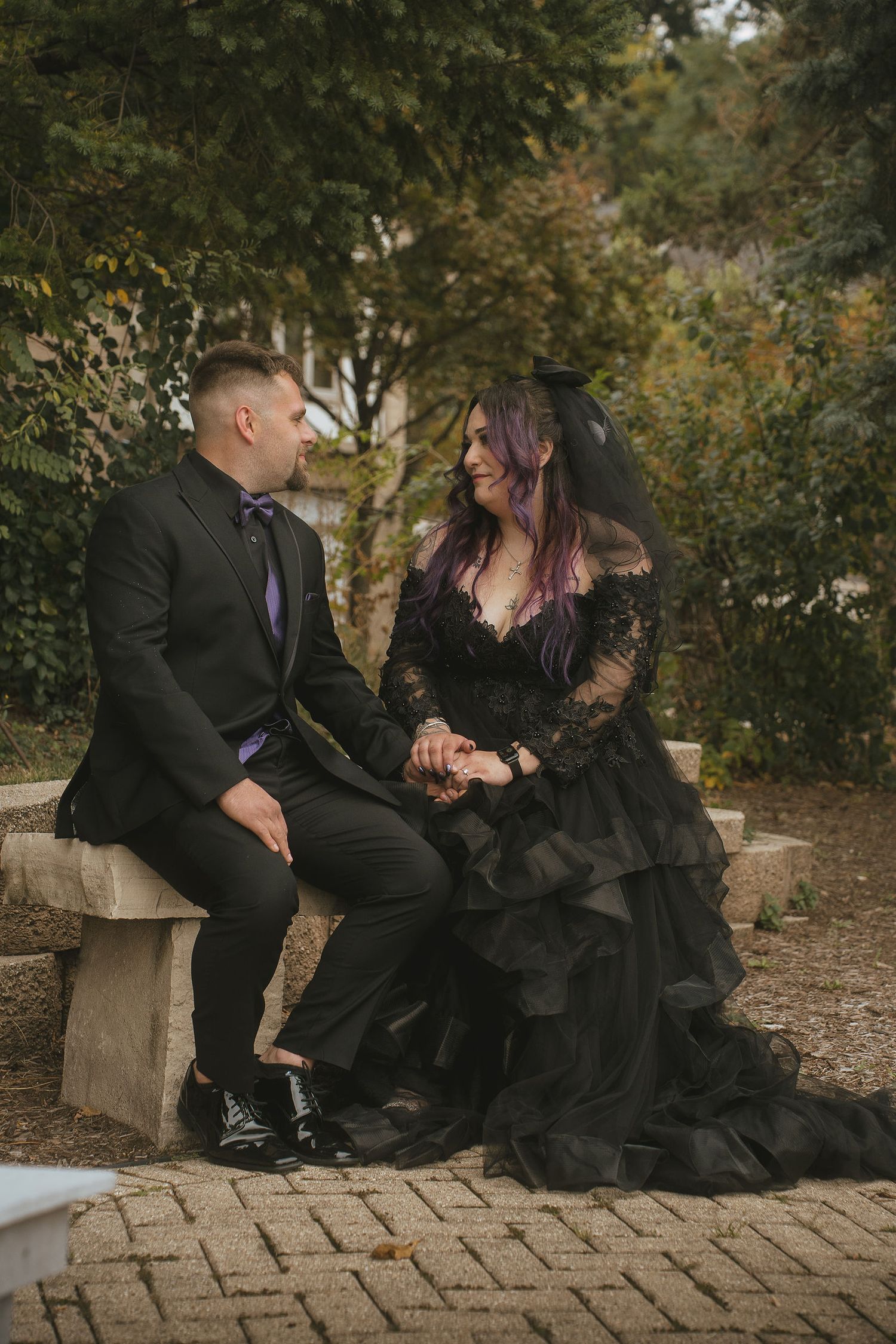 A couple in formal black attire share intimate moments while sitting on stone steps in an autumn garden setting.