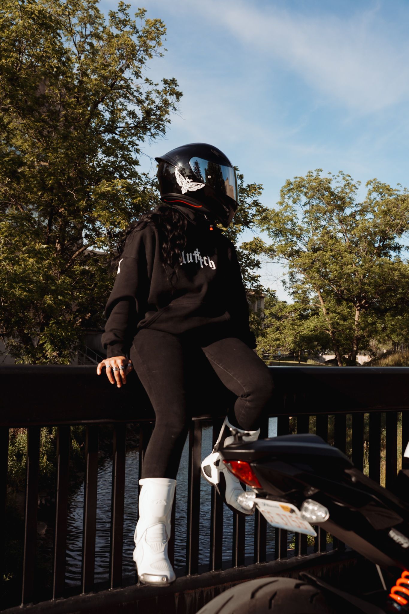 Person in black outfit and helmet sitting on motorcycle railing bridge with trees background.