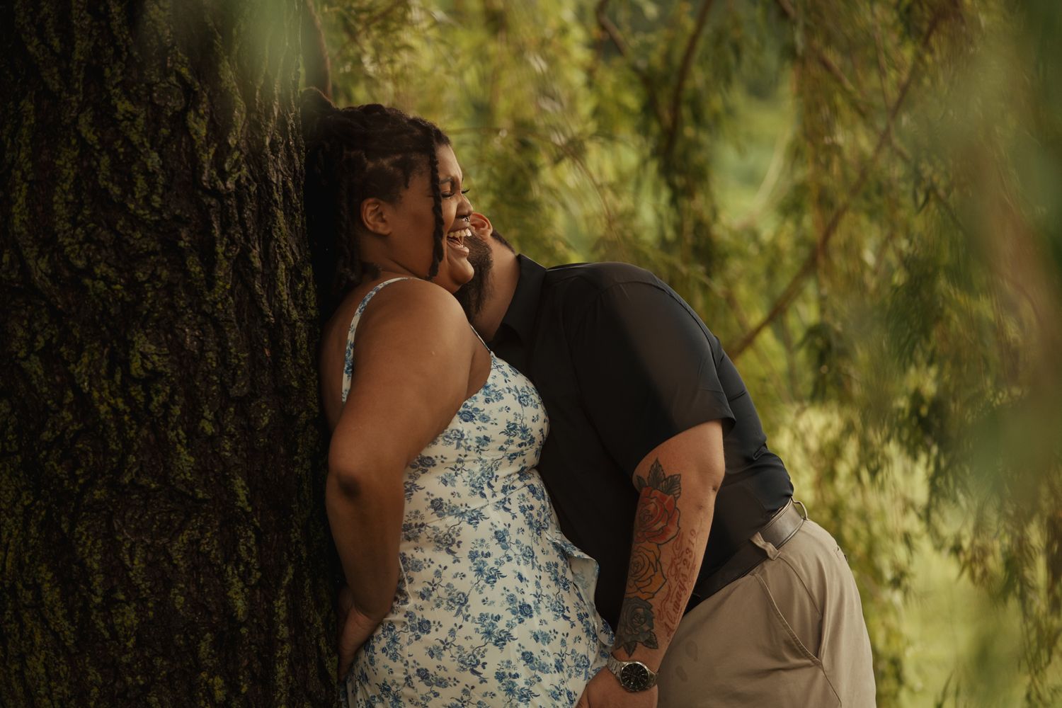 Couple sharing a romantic moment leaning against a large tree outdoors, surrounded by lush green foliage.