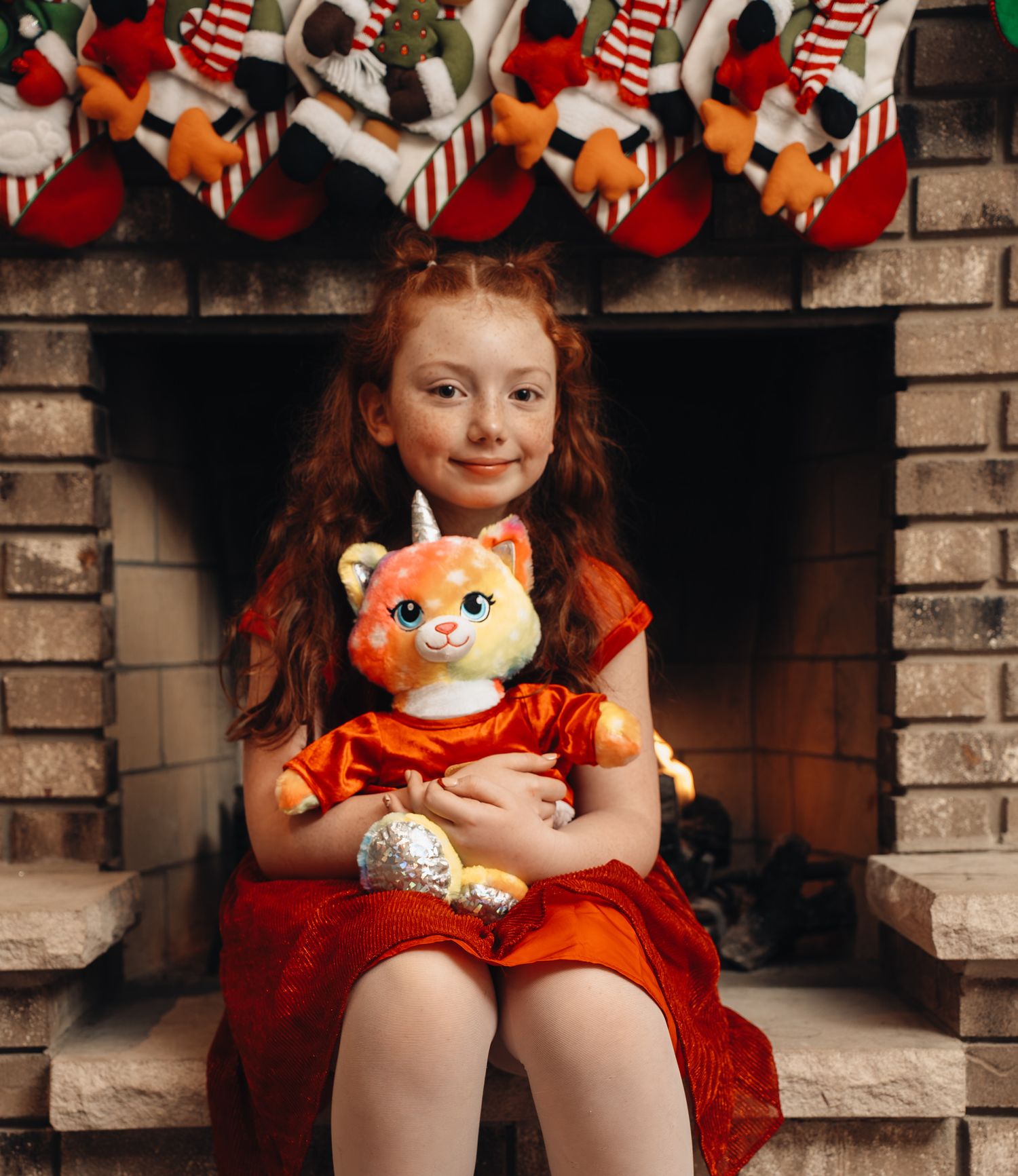 Child in orange dress sits on brick hearth with plush fox toy in cozy holiday setting.