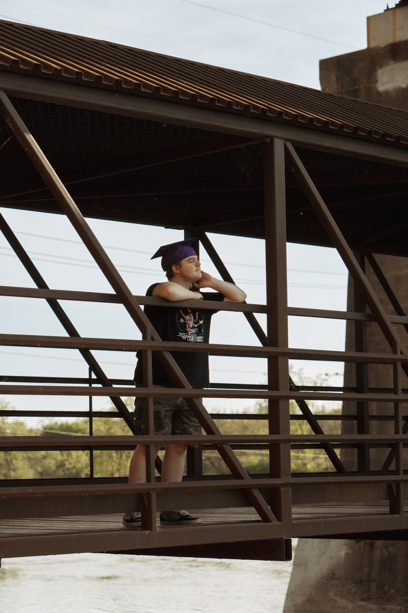 Young man in black t-shirt and purple cap standing on a metal bridge railing, gazing into the distance.