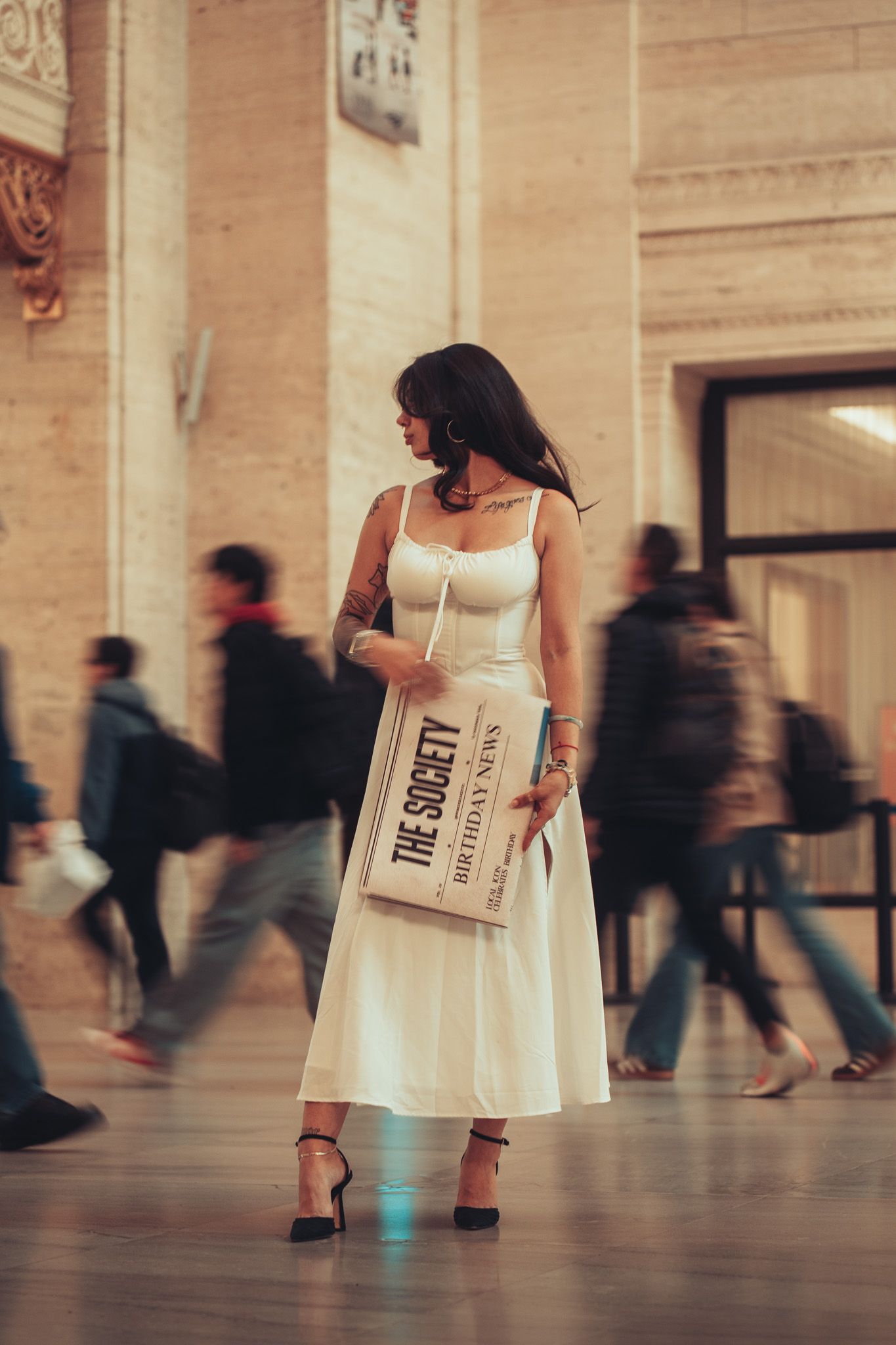 A woman in a white dress holds a shopping bag while standing in a busy hallway with blurred people walking past.