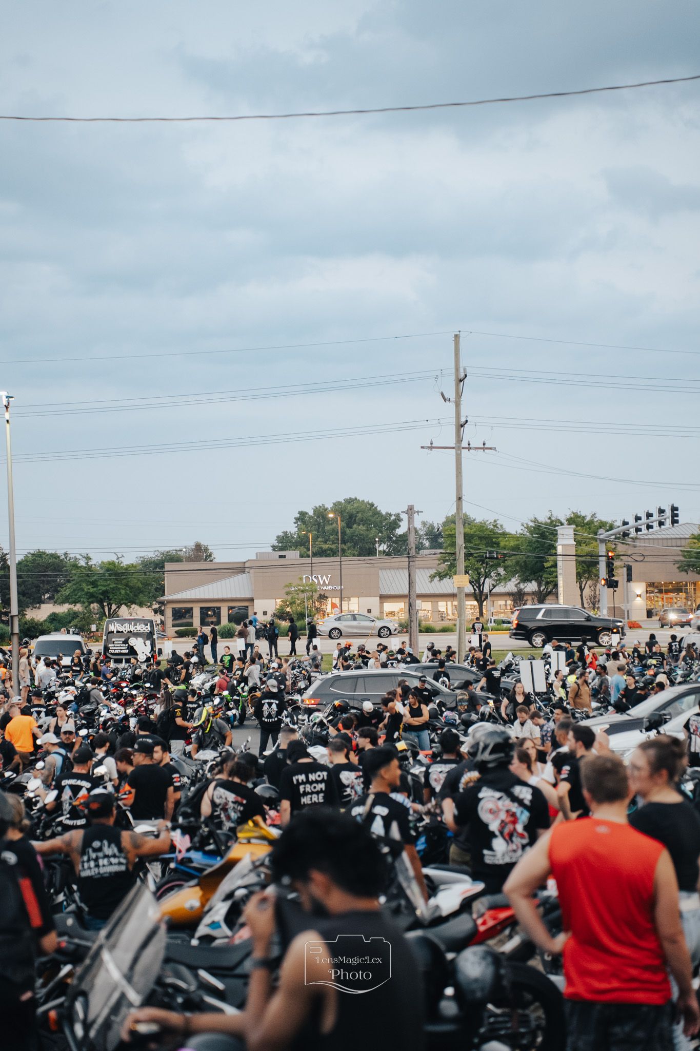 A large crowd of motorcycles and riders gathered in a parking lot under power lines and cloudy skies.