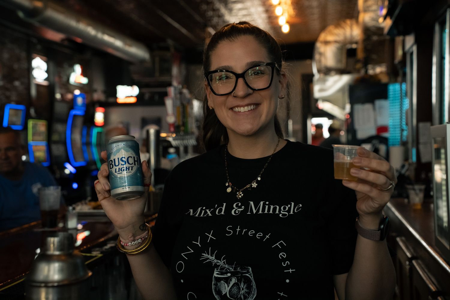 A bartender holds up a Coors Light can and shot glass while working behind a dimly lit bar counter.