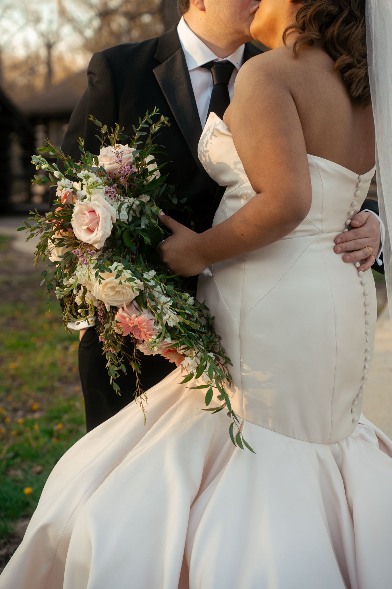 Bride in strapless mermaid gown holding lush floral bouquet, kissing groom in black tuxedo outdoors.