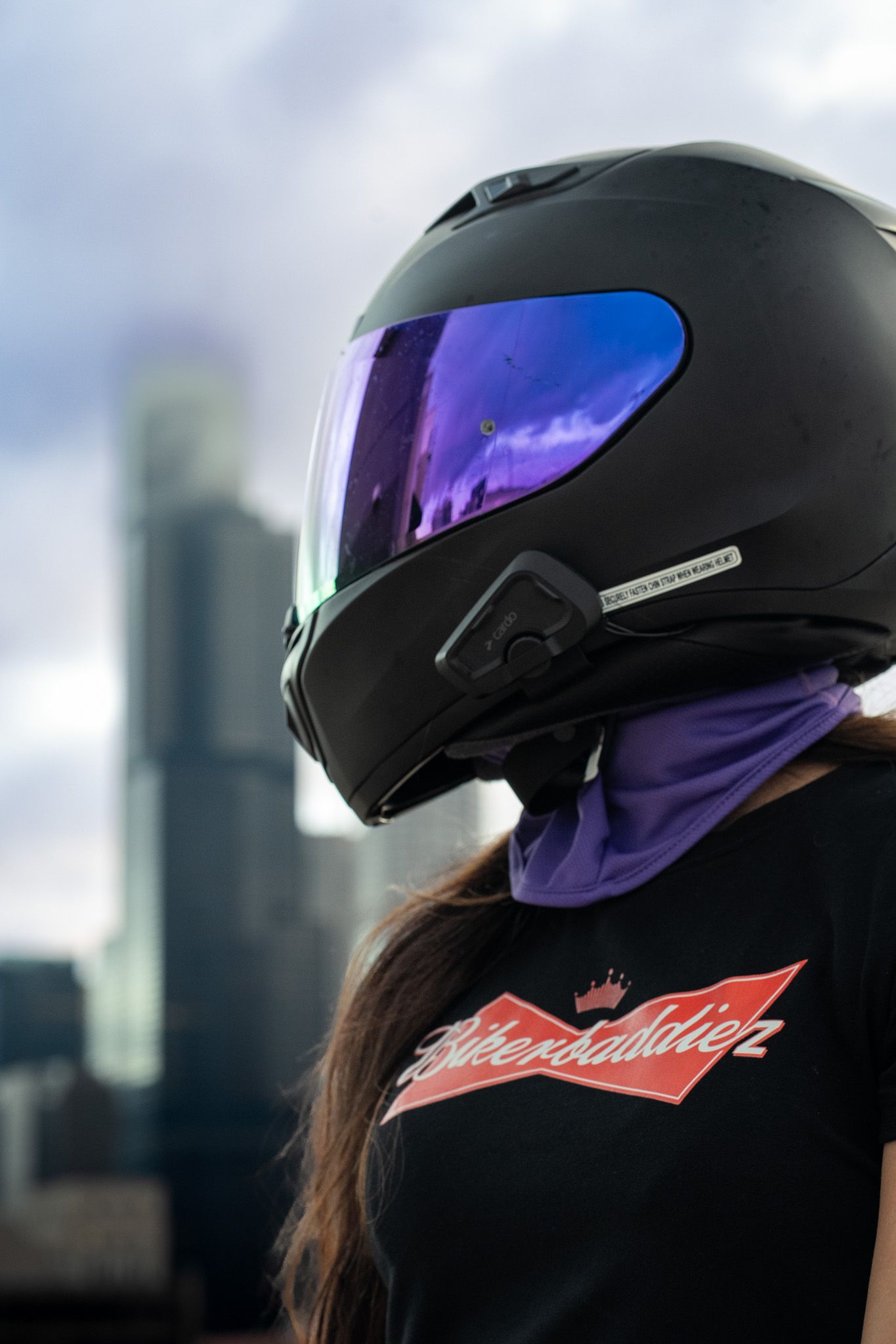A black motorcycle helmet with iridescent visor reflects the urban skyline behind it.
