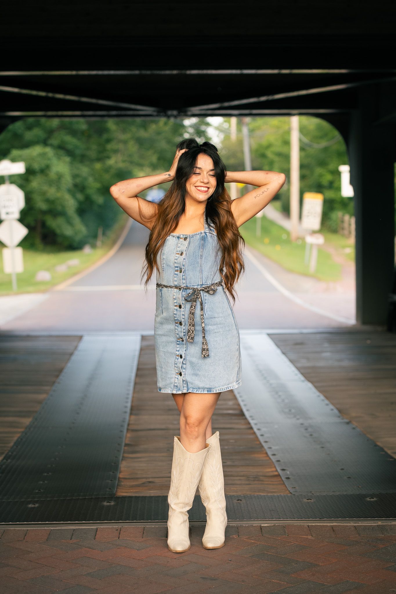 A person in denim and white boots poses on a brick walkway under an overpass.