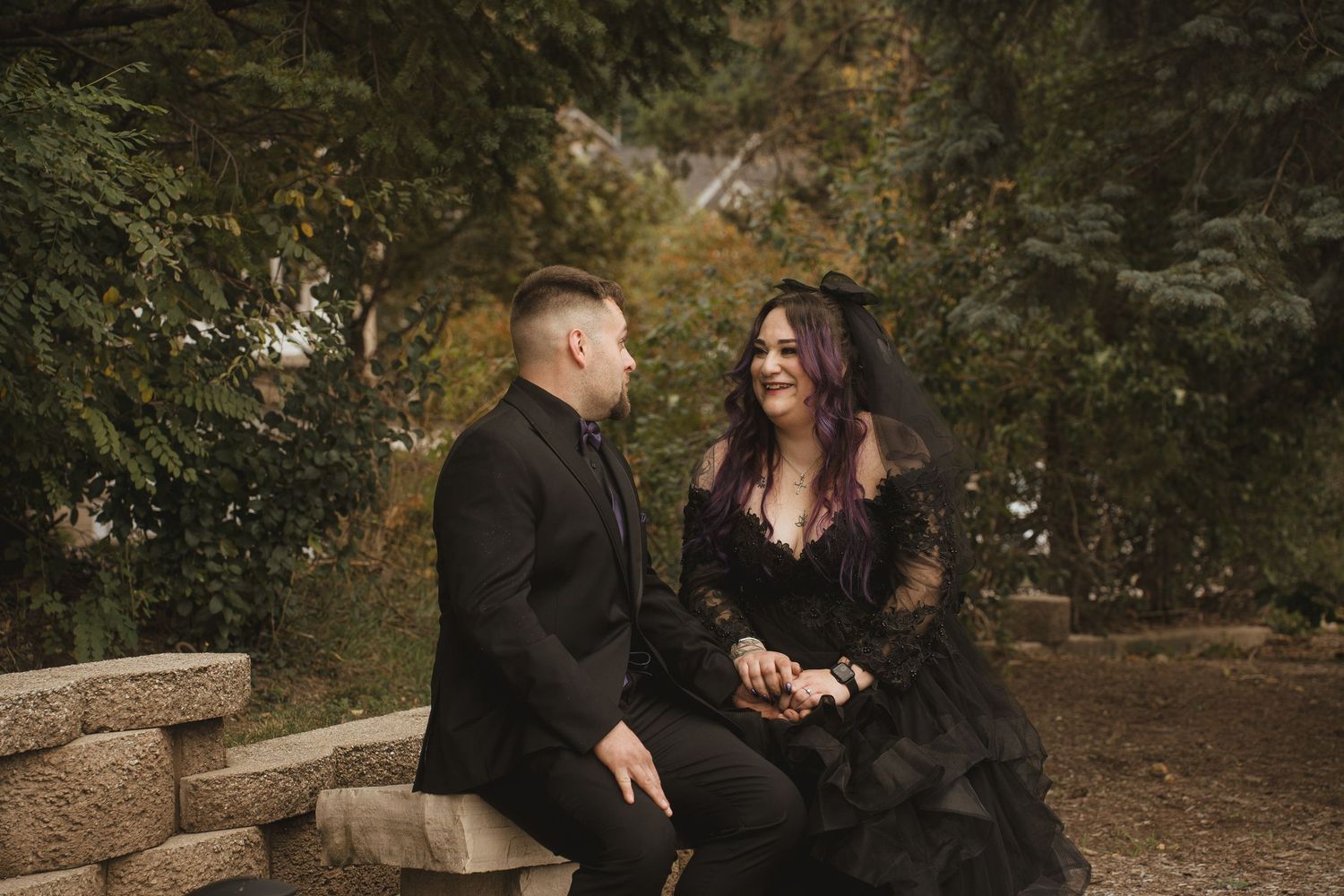 Gothic couple shares tender moment on stone bench surrounded by fall foliage.