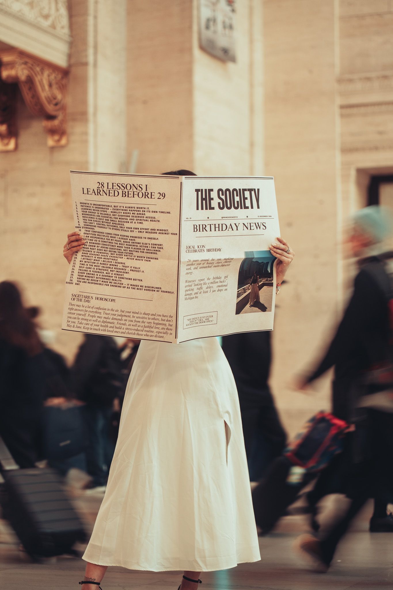 Person in white dress holding newspapers while others walk past in blurred motion at indoor venue.