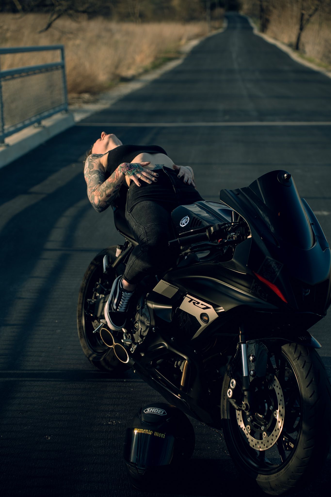 Tattooed man leaning over a black Yamaha R7 sport motorcycle on an empty road, wearing all black attire.