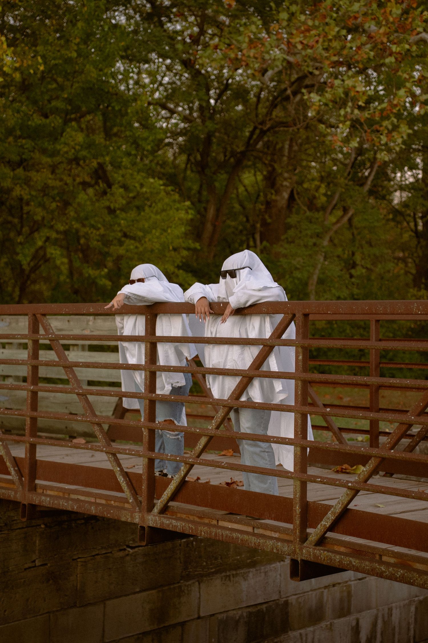 Ghost-costumed figures in white sheets stand on a wooden bridge in an autumn setting.