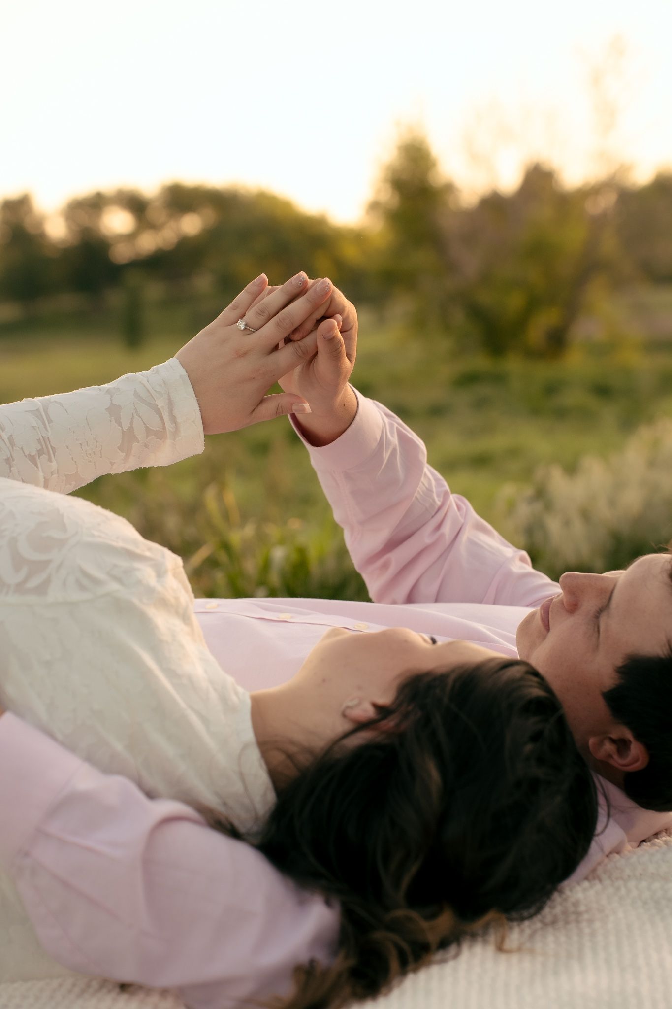 Couple lying on blanket outdoors holding hands at sunset, wearing white and pink, with green nature background.
