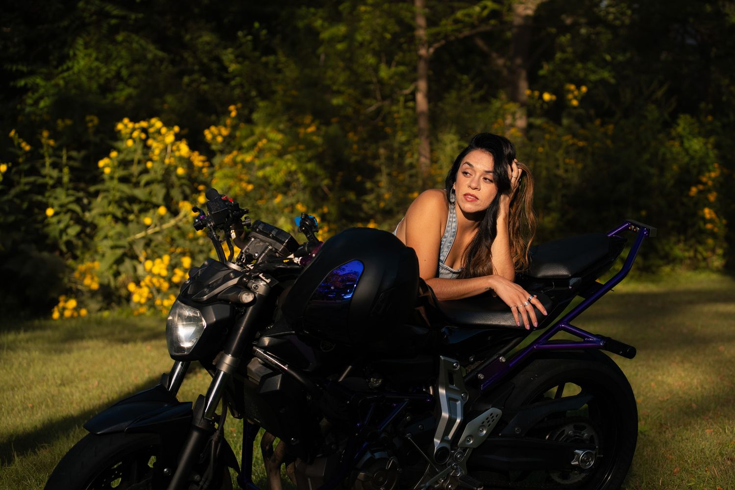 A person reclines on a black motorcycle in an outdoor setting with yellow flowers.