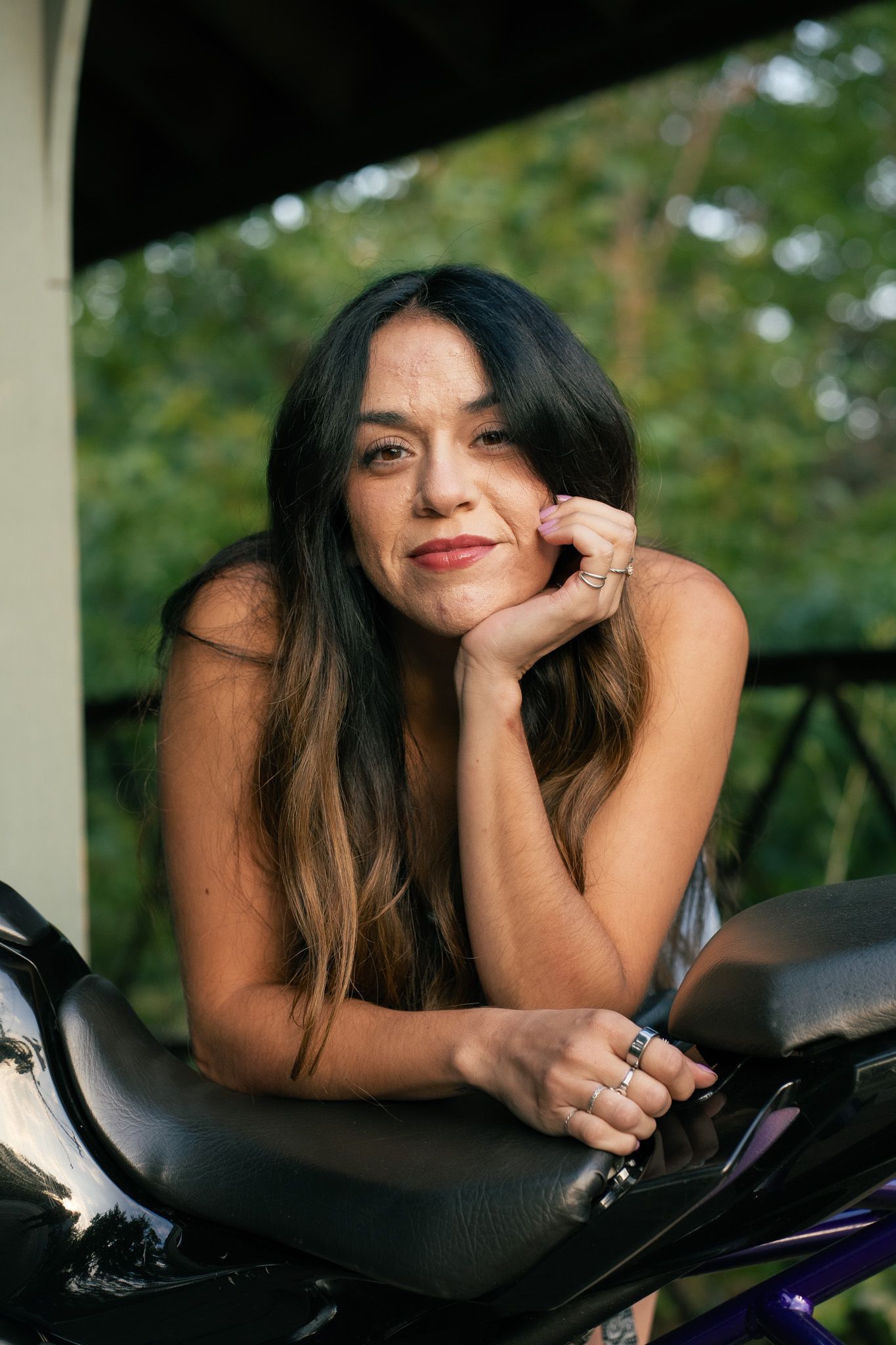 A person in dark clothing poses on a motorcycle in an outdoor setting with green foliage in the background.