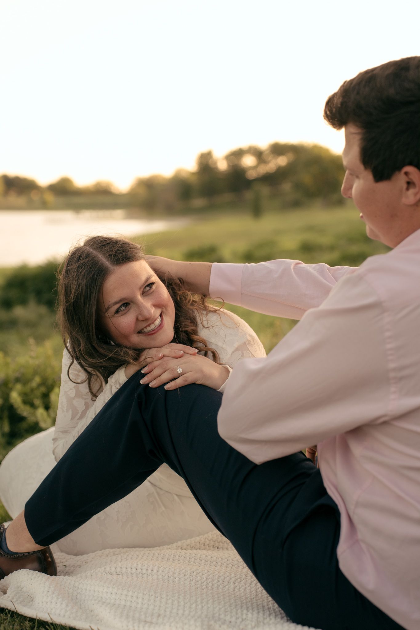 Couple sitting on blanket by lake at sunset, woman smiling up at man in pink shirt during romantic engagement photo.