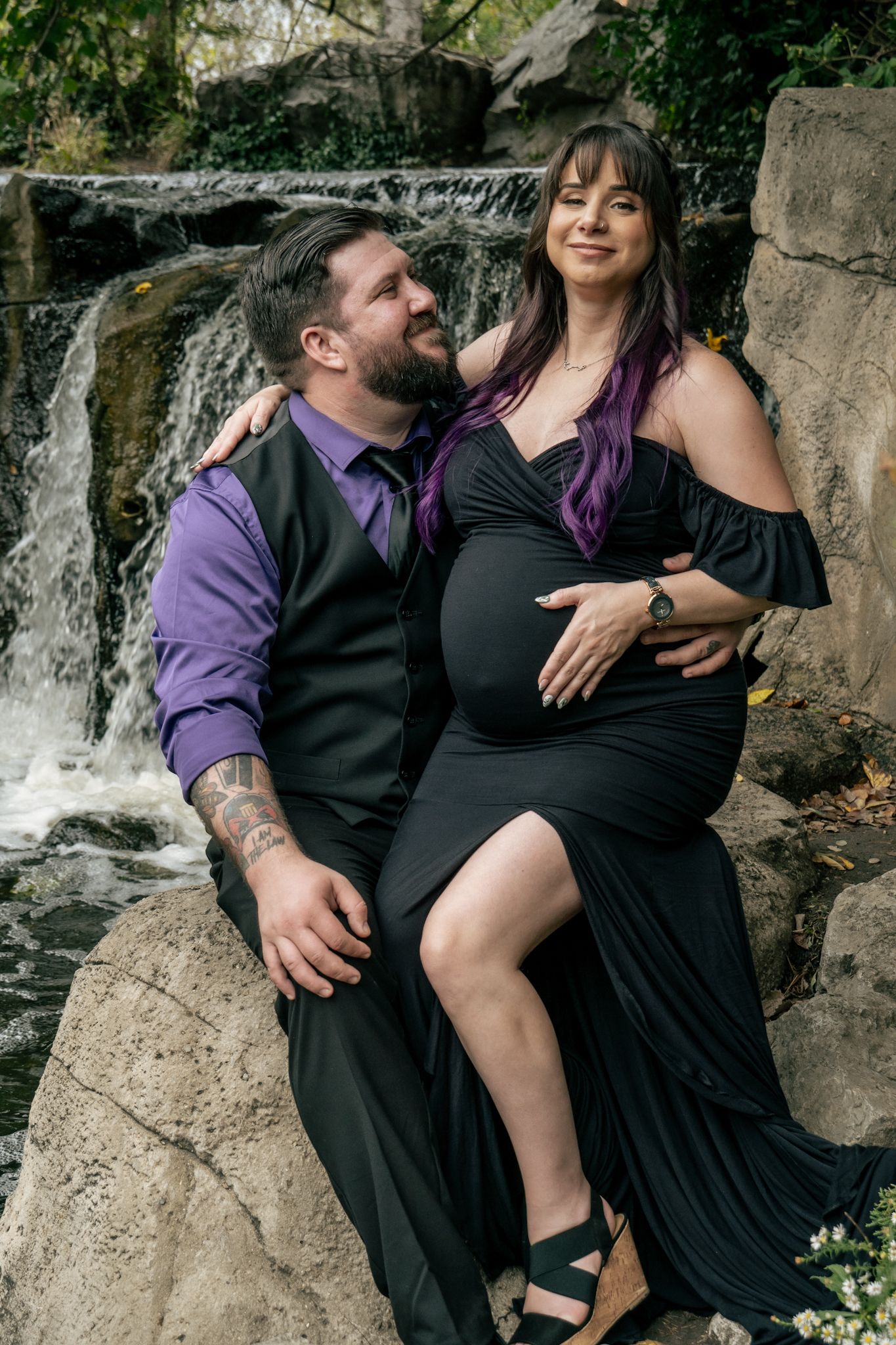 Couple in coordinated black and purple formal wear pose intimately by a scenic waterfall for romantic photos.