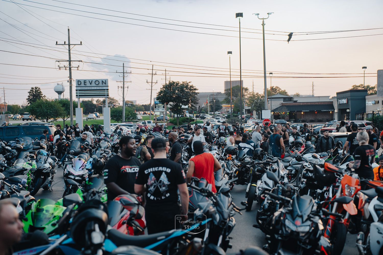 Large group of motorcycles and riders gathered at dusk in a parking lot for a motorcycle meetup event.