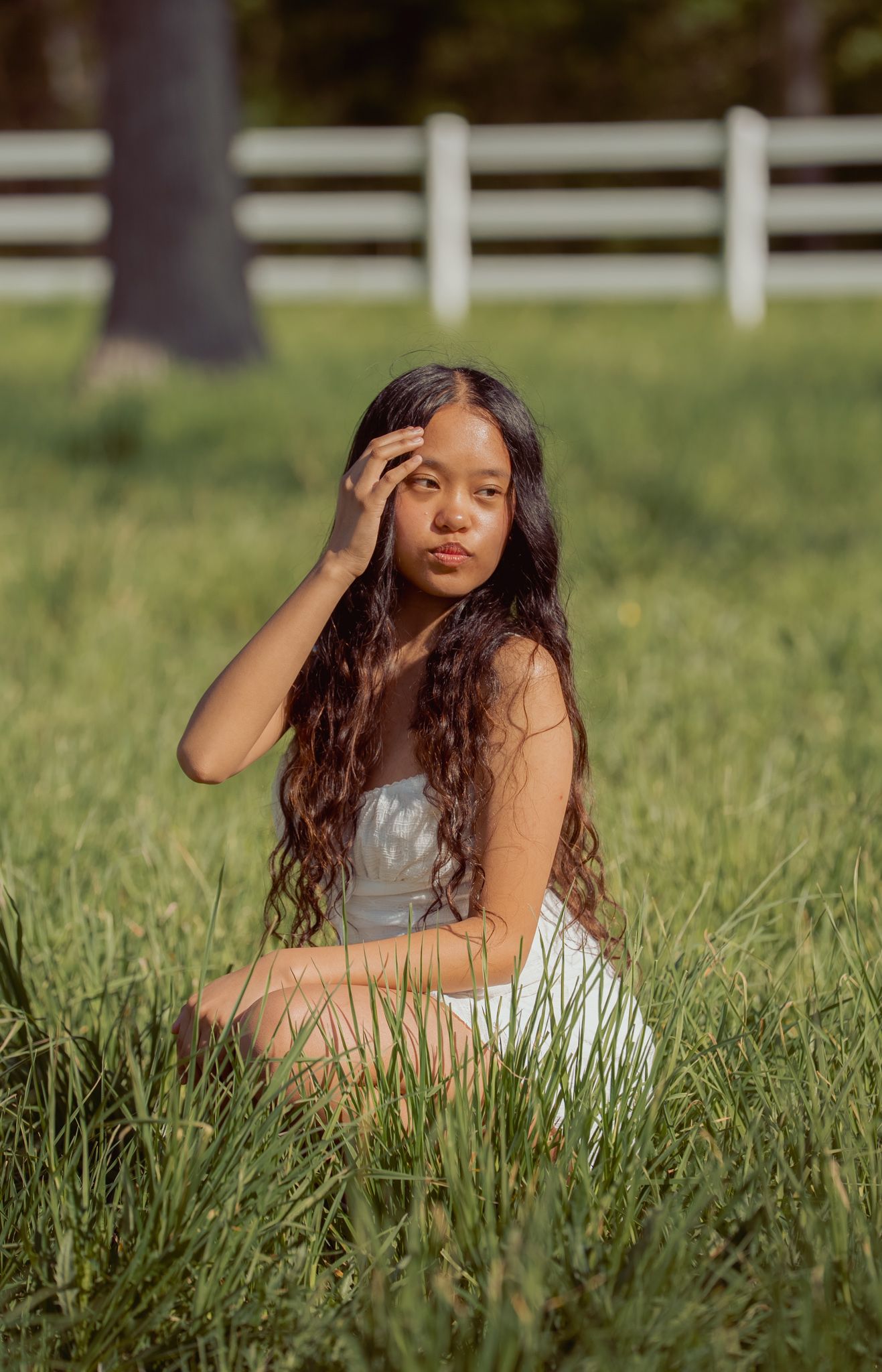 Young woman with long curly hair sits in green grass wearing a white dress at a farm.